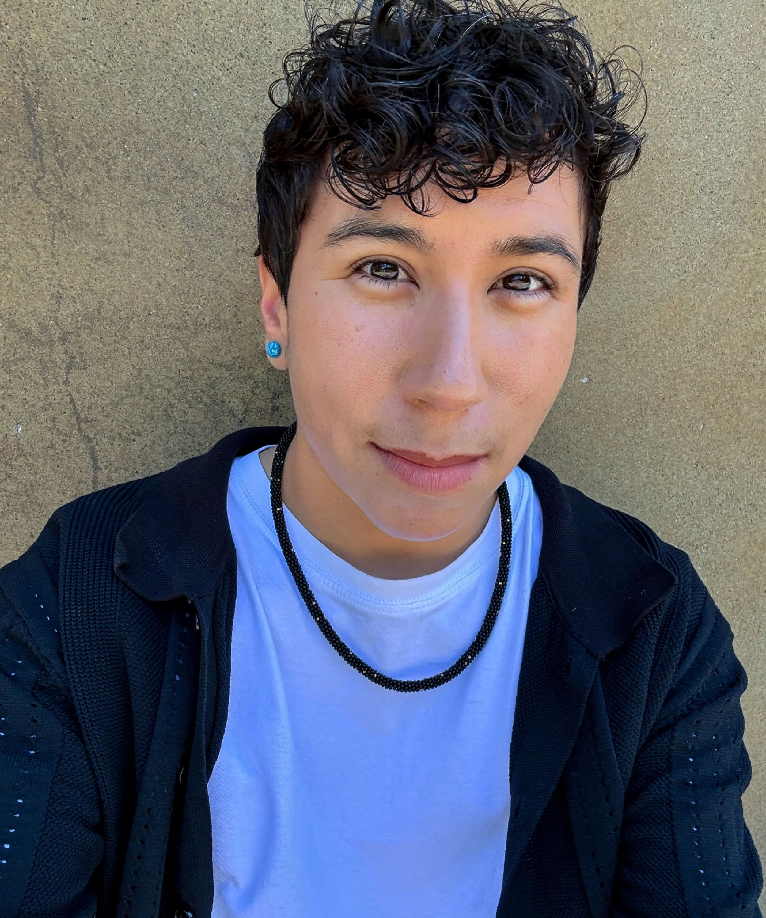 A person with short, curly black hair and light brown skin, wearing a white t-shirt, a black jacket, blue circular earrings, and a beaded necklace, standing against a plain beige wall.