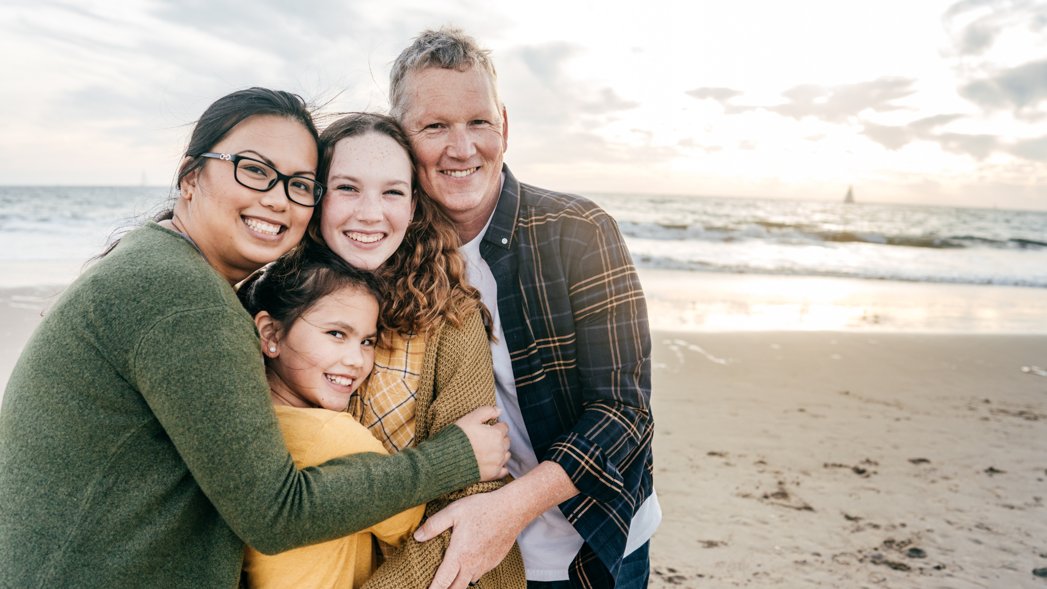 A family of four hugging and smiling on the beach at sunset, with the ocean in the background.