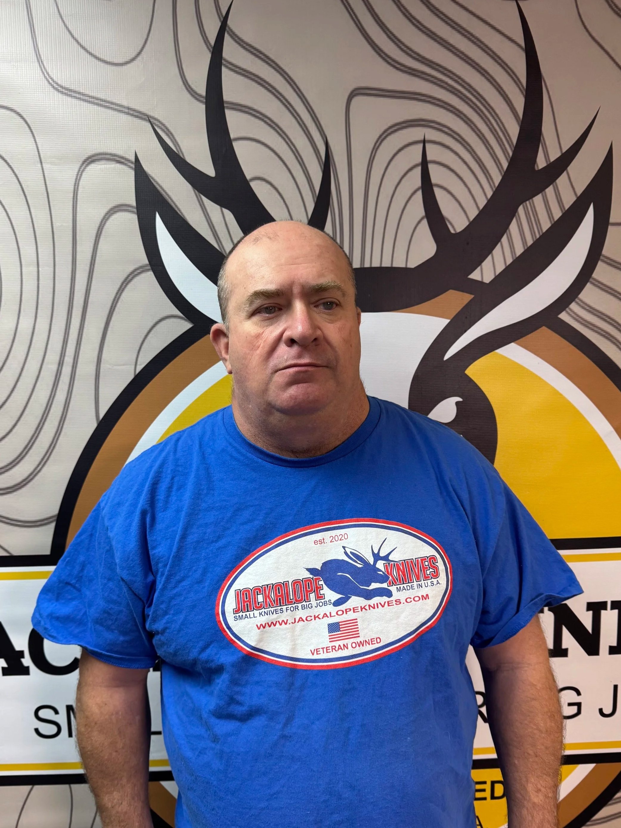 A man with a serious expression wearing a blue T-shirt in front of a logo featuring a stylized deer head and the text 'JACKALOPE KNIVES'.