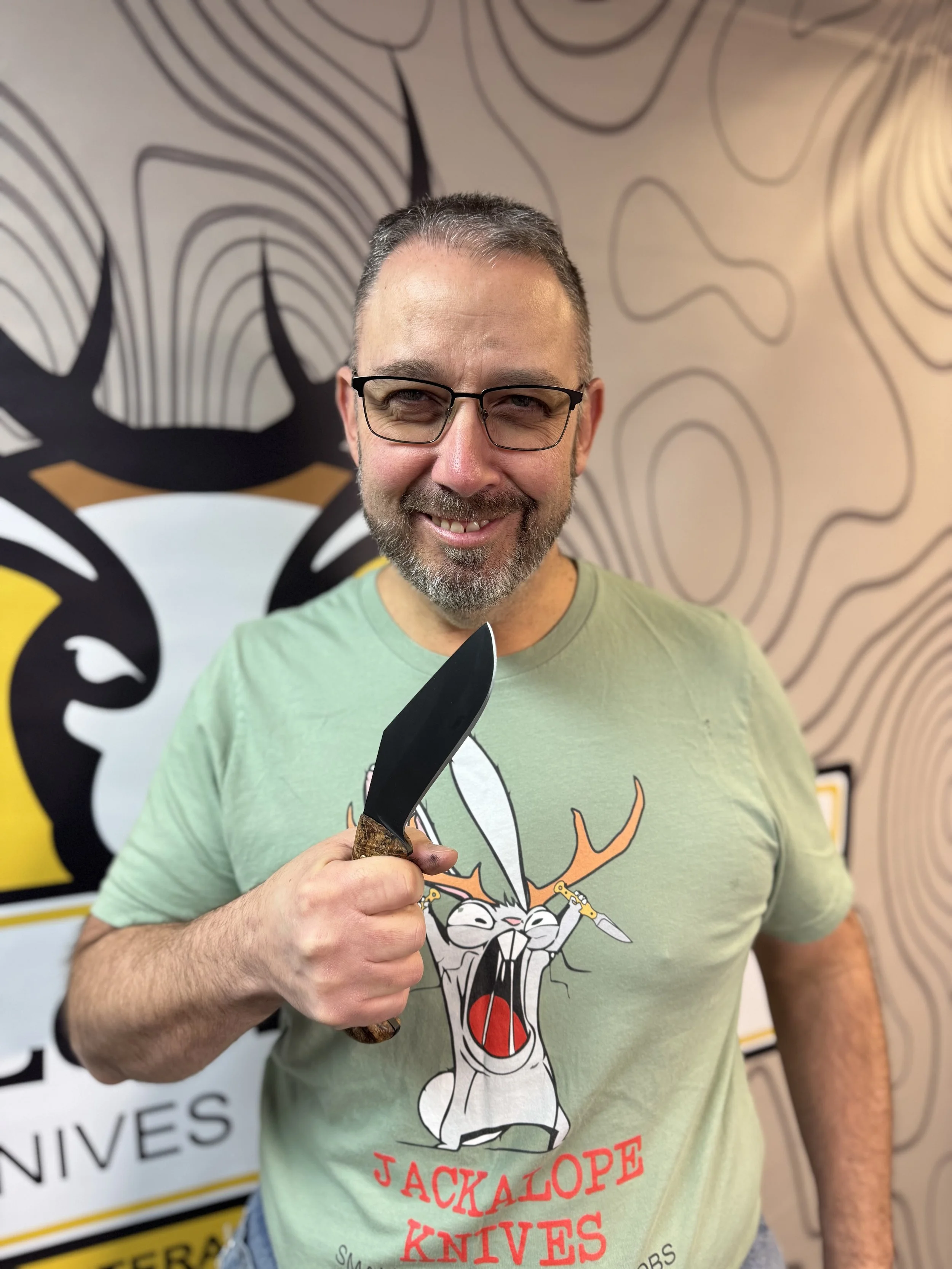 A man with glasses and a beard holding a kitchen knife, wearing a light green T-shirt with a cartoon rabbit with antlers and the text "Jackalope Knives".