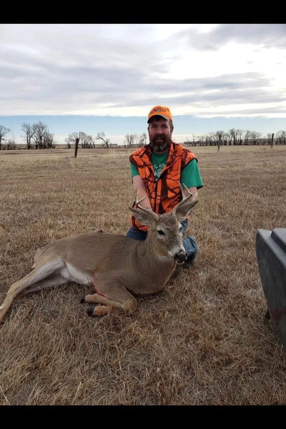 A man is kneeling on grass in an open field, holding a large deer with antlers. The man is wearing an orange hunting hat and vest, with a green shirt underneath. The field has a few scattered trees and a cloudy sky above.