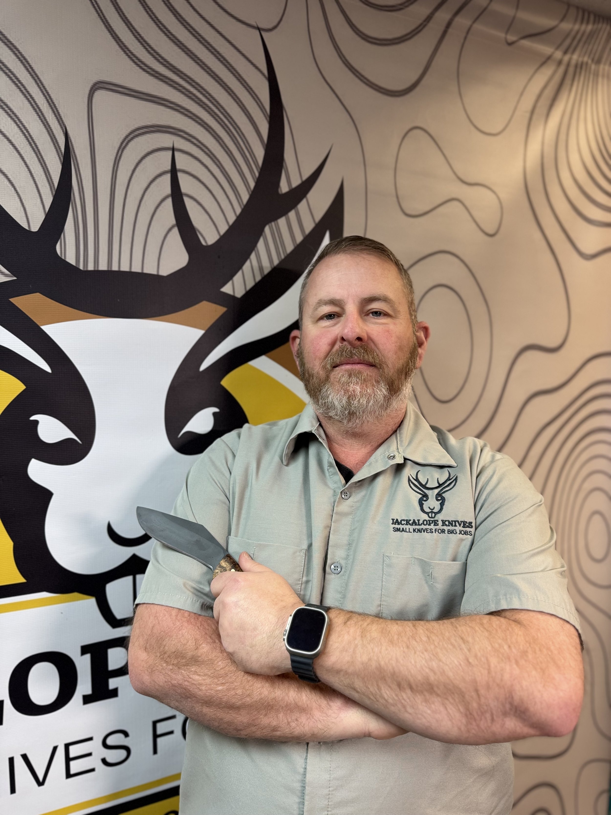 A man with a beard wearing a blurred smartwatch, holding a knife, standing in front of a wall with a deer logo and a wooden pattern background, wearing a khaki shirt with a deer logo and the text "JACKALOPE KNIVES SMALL KNIVES FOR BIG JOBS."