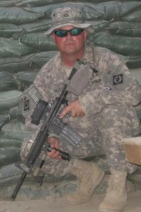 A soldier in camouflage uniform kneels with a rifle in front of sandbags, wearing sunglasses and a boonie hat.
