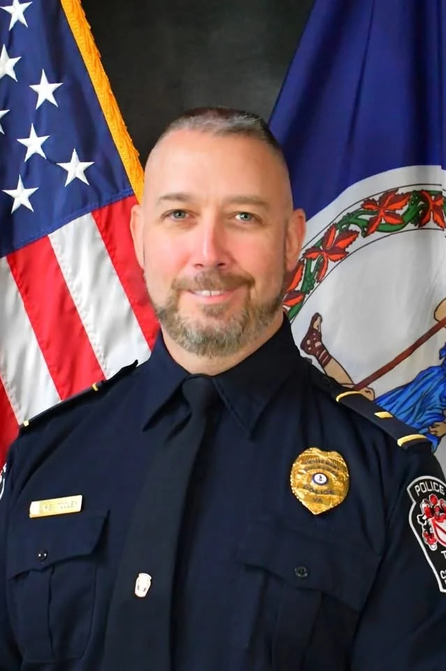 A male police officer with a beard and short hair, standing in front of the American flag and a state flag, wearing a dark police uniform with a badge and patches.