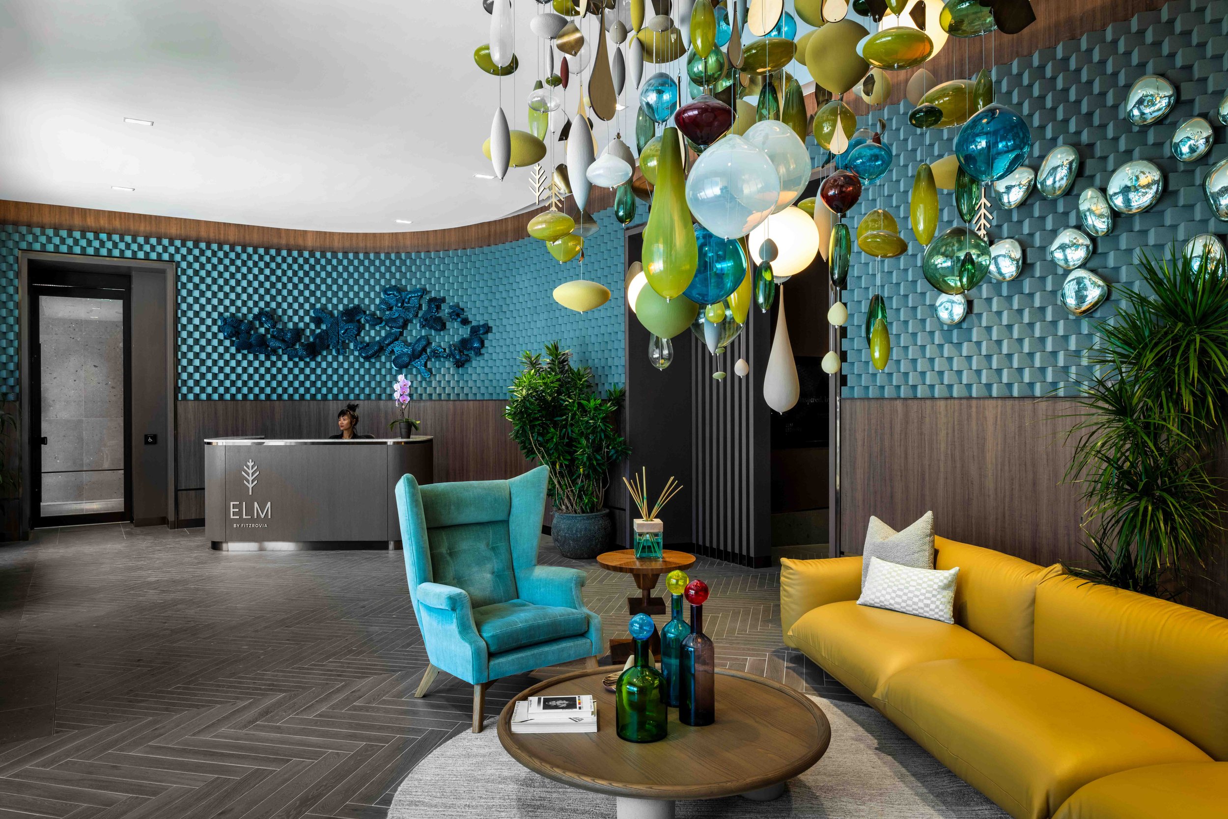 Modern hotel lobby with colorful hanging glass sculpture, blue and yellow seating, reception desk, and decorative wall art.