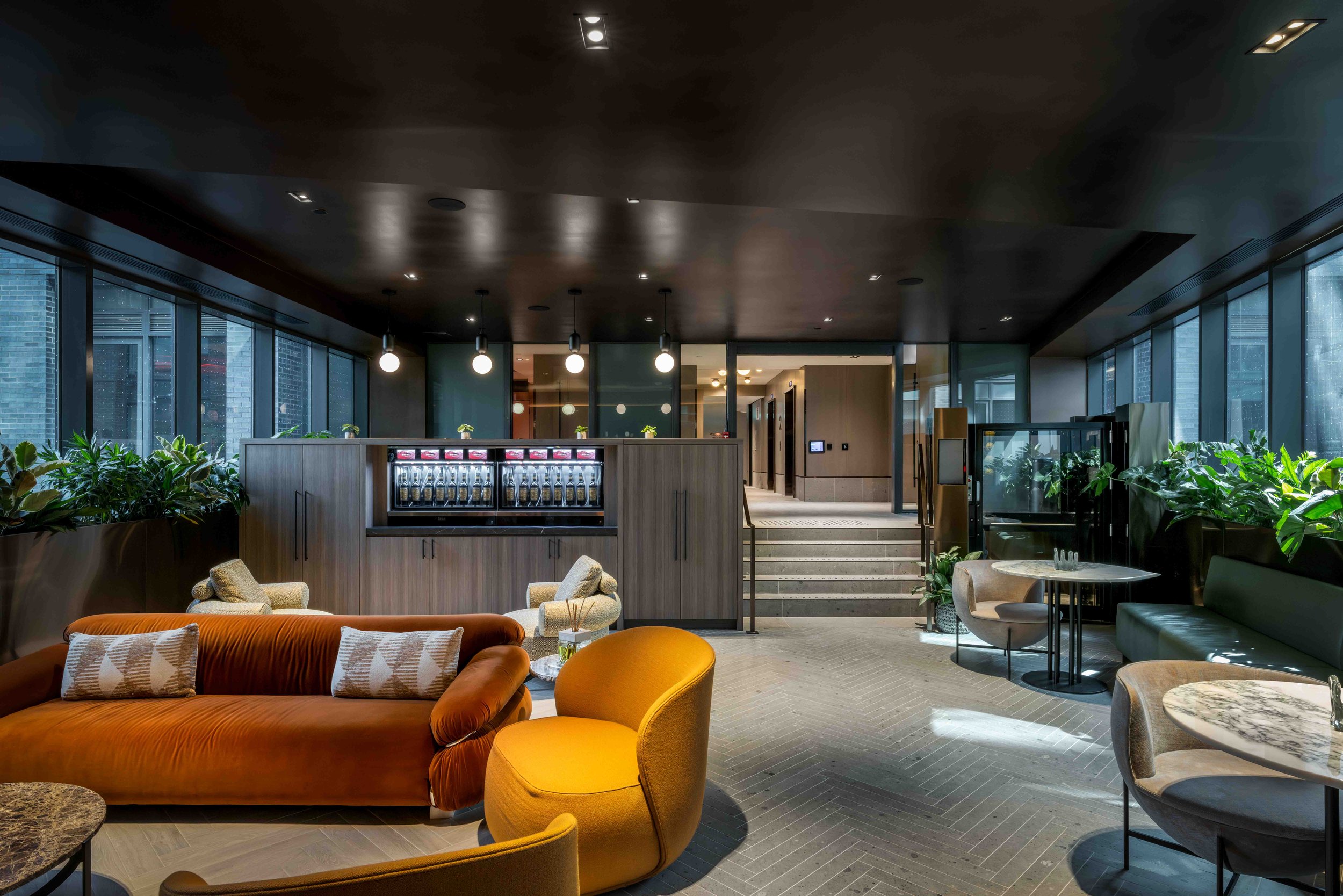 Modern lounge area with orange sofa, round chairs, and marble tables, featuring large windows and potted plants, illuminated by pendant lights.