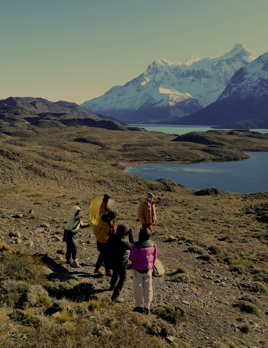 Group of hikers walking on a mountain trail with landscapes of lakes, hills, and snow-capped mountains in the background.