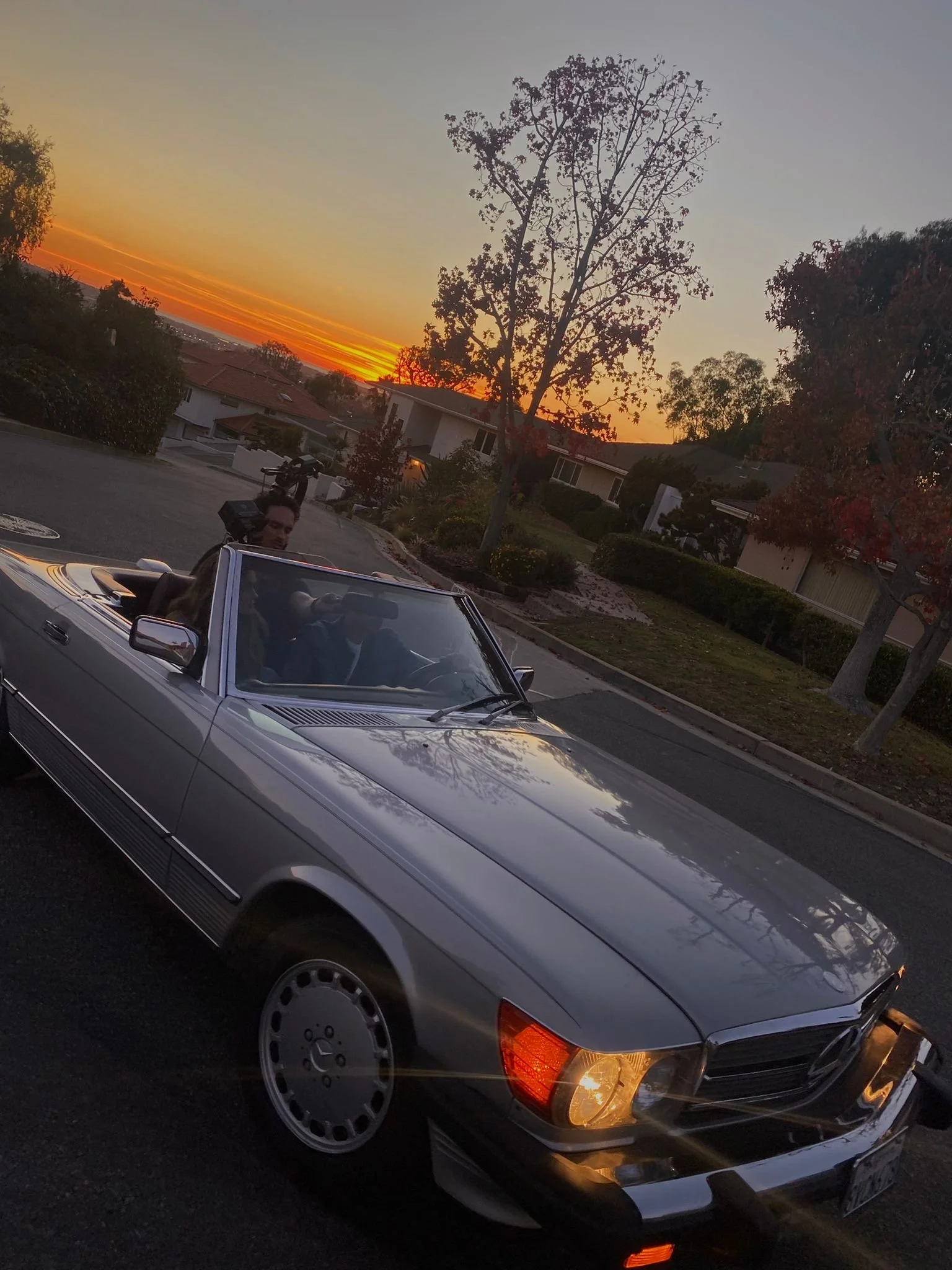 Three people in a convertible car on a street at sunset. One person, standing, holding a camera; two others seated inside, smiling and making gestures.