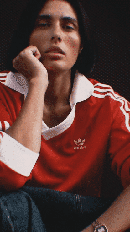 Person with dark hair wearing a red Adidas sports jacket with white stripes, sitting with one hand resting on their face, against a dark background.