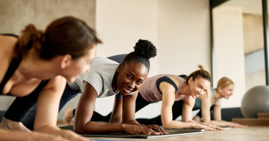 A group of women doing forearm planks on exercise mats in a workout studio. They are looking at each other and smiling.