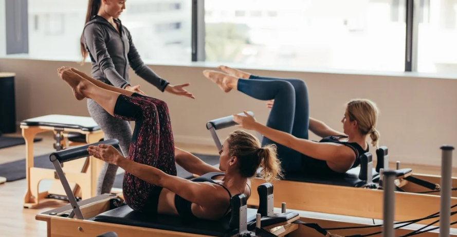 Two women perform Pilates on reformers while an instructor guides them in a bright studio with large windows.