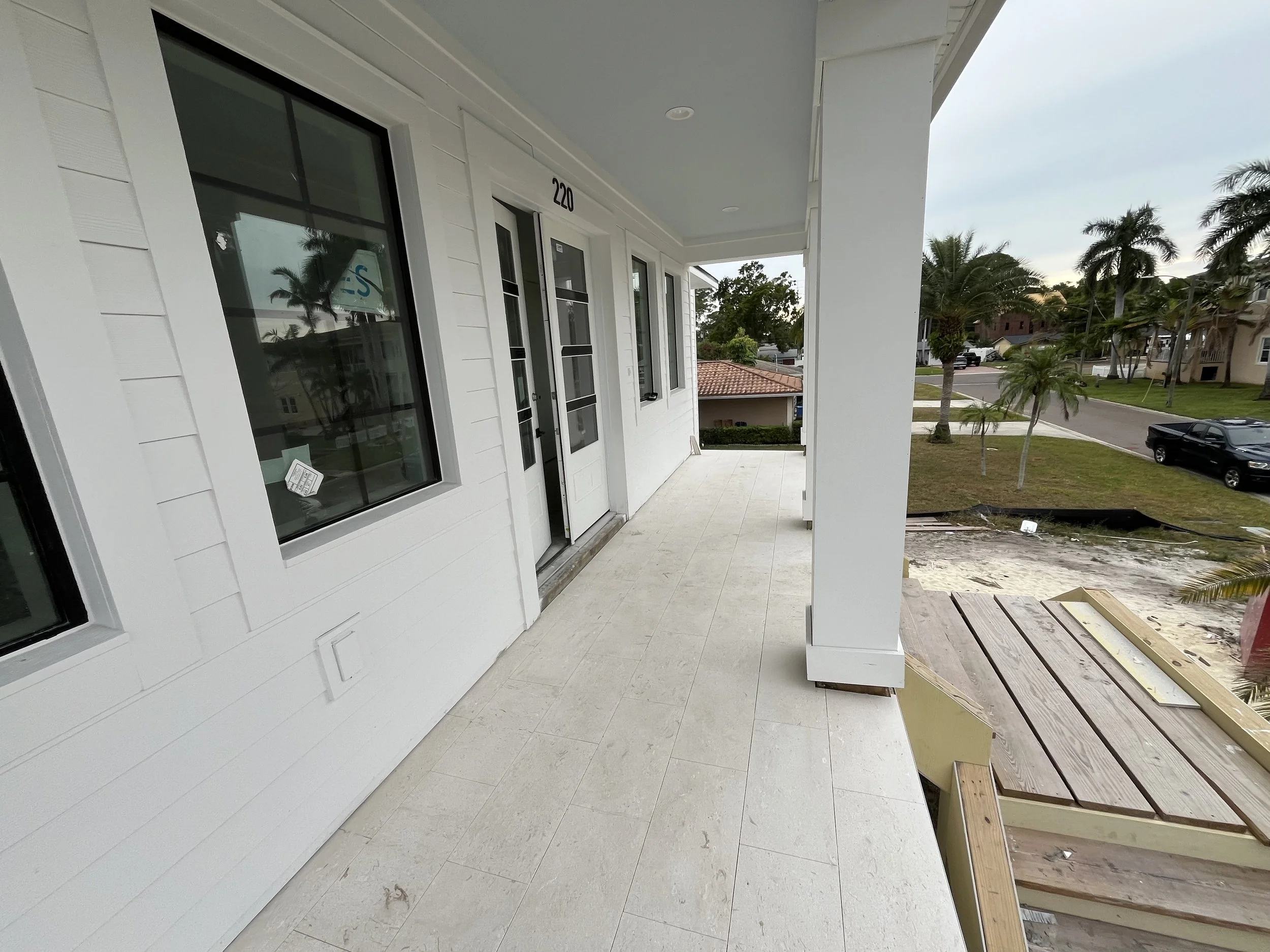 A newly constructed white house balcony with beige tile flooring, several large windows, and a door. The balcony overlooks a street with palm trees and parked cars, and there is a construction site with unfinished wooden frames on the right side.