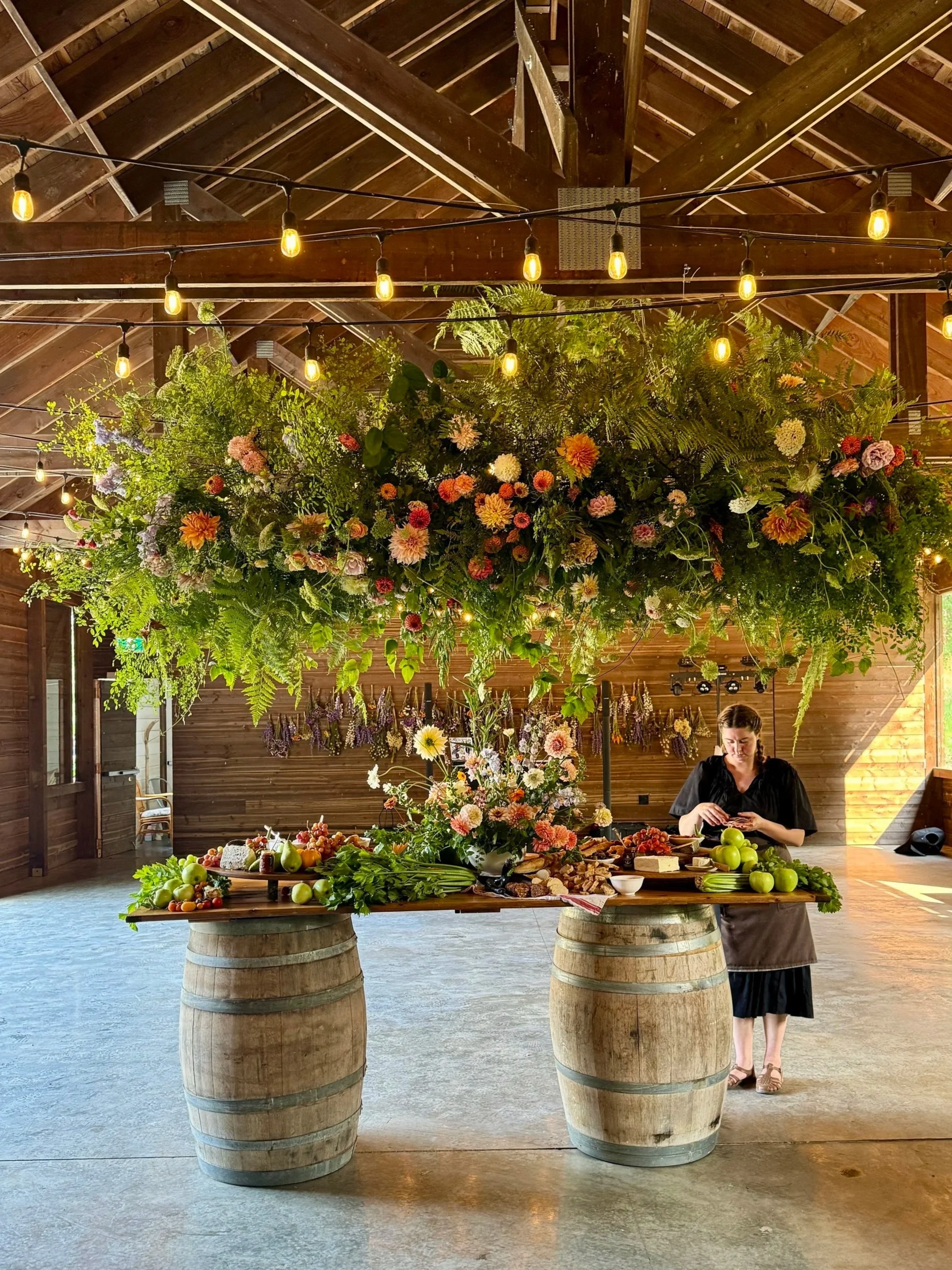 A woman at a table with a large floral arrangement and food, standing inside a barn with a high, wooden ceiling and string lights hanging overhead.