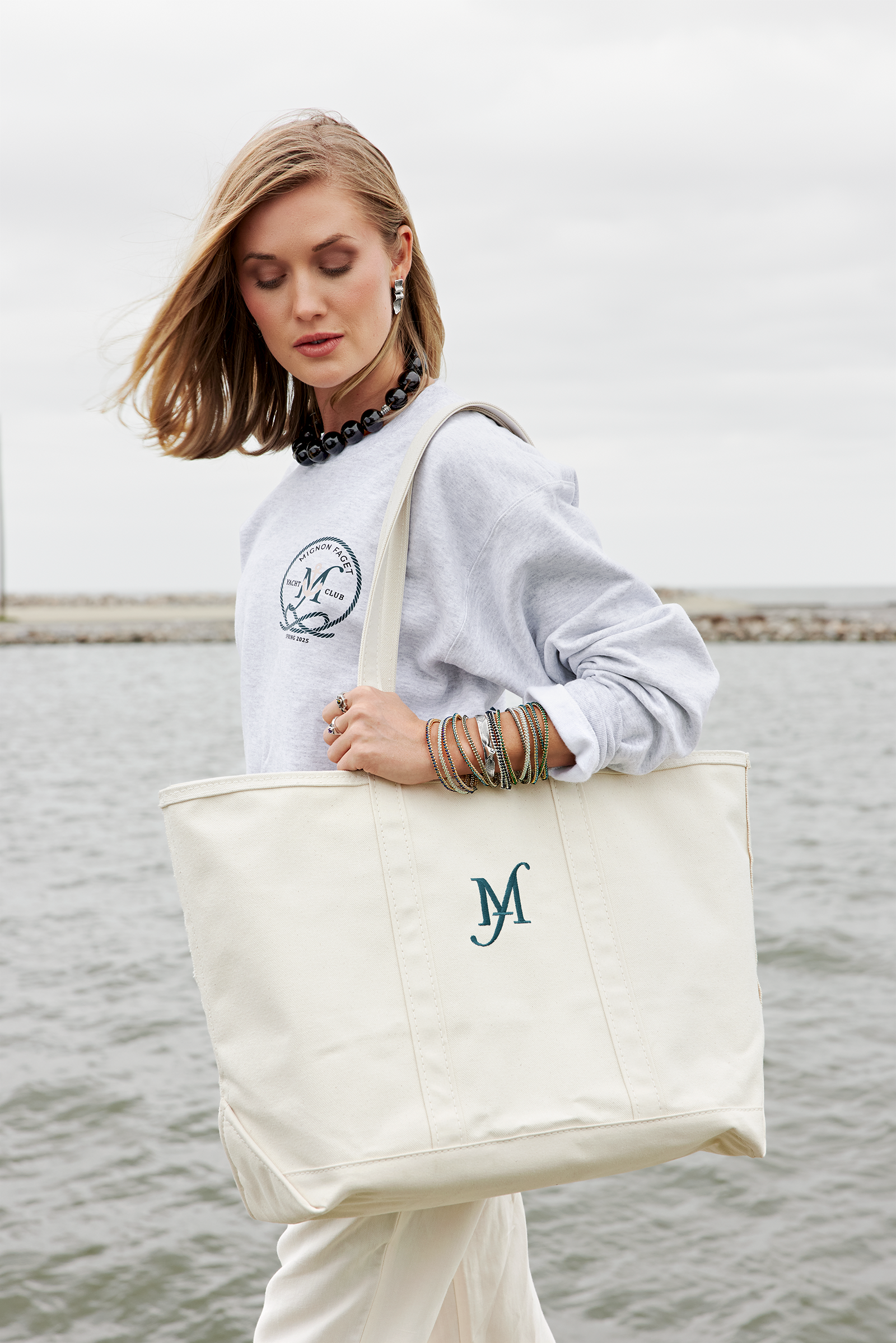 A woman with shoulder-length blonde hair wearing a gray sweatshirt, a chunky beaded necklace, and multiple bracelets, standing outdoors near a body of water, holding a large beige tote bag with initials 'M' and 'A' embroidered on it.