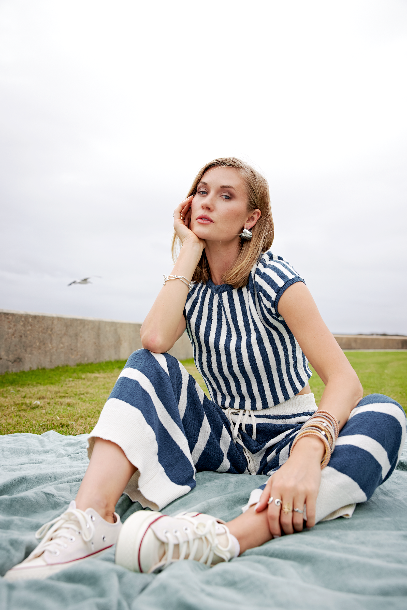 Young woman sitting on a blanket outdoors, wearing a blue and white striped outfit, earrings, and bracelets, with a cloudy sky and green grass in the background.
