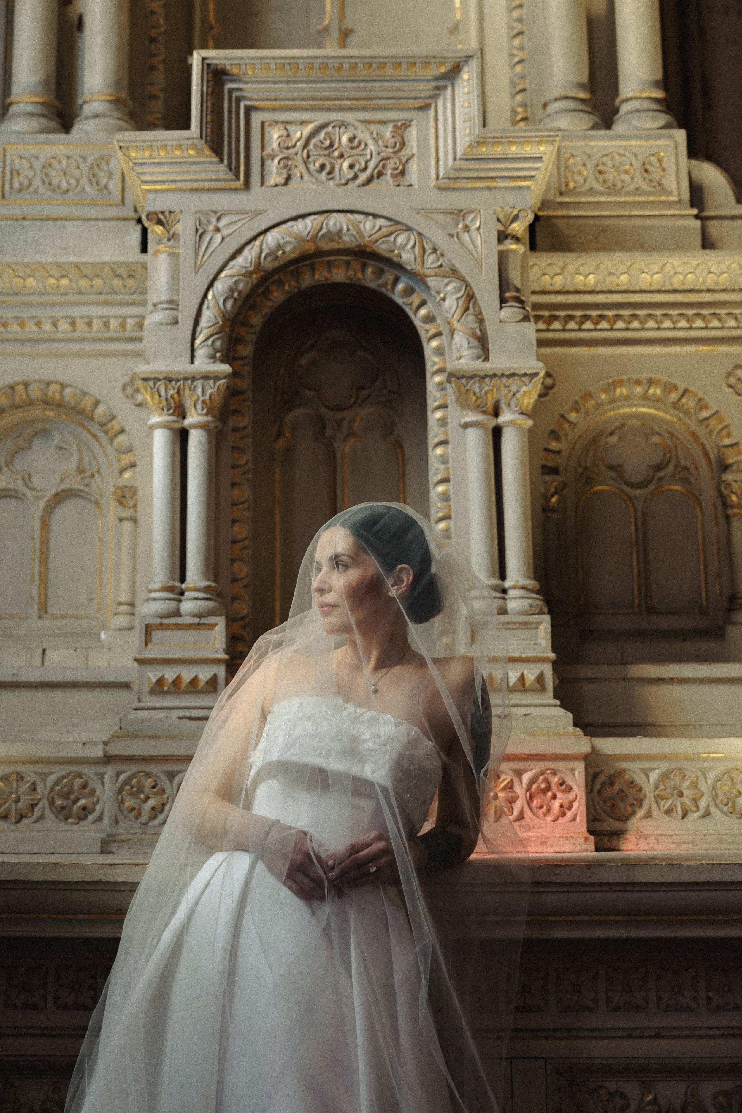A bride in a white wedding dress with a veil stands in front of an ornate, gold-accented marble altar inside a church, turned slightly to her left with her hands clasped.