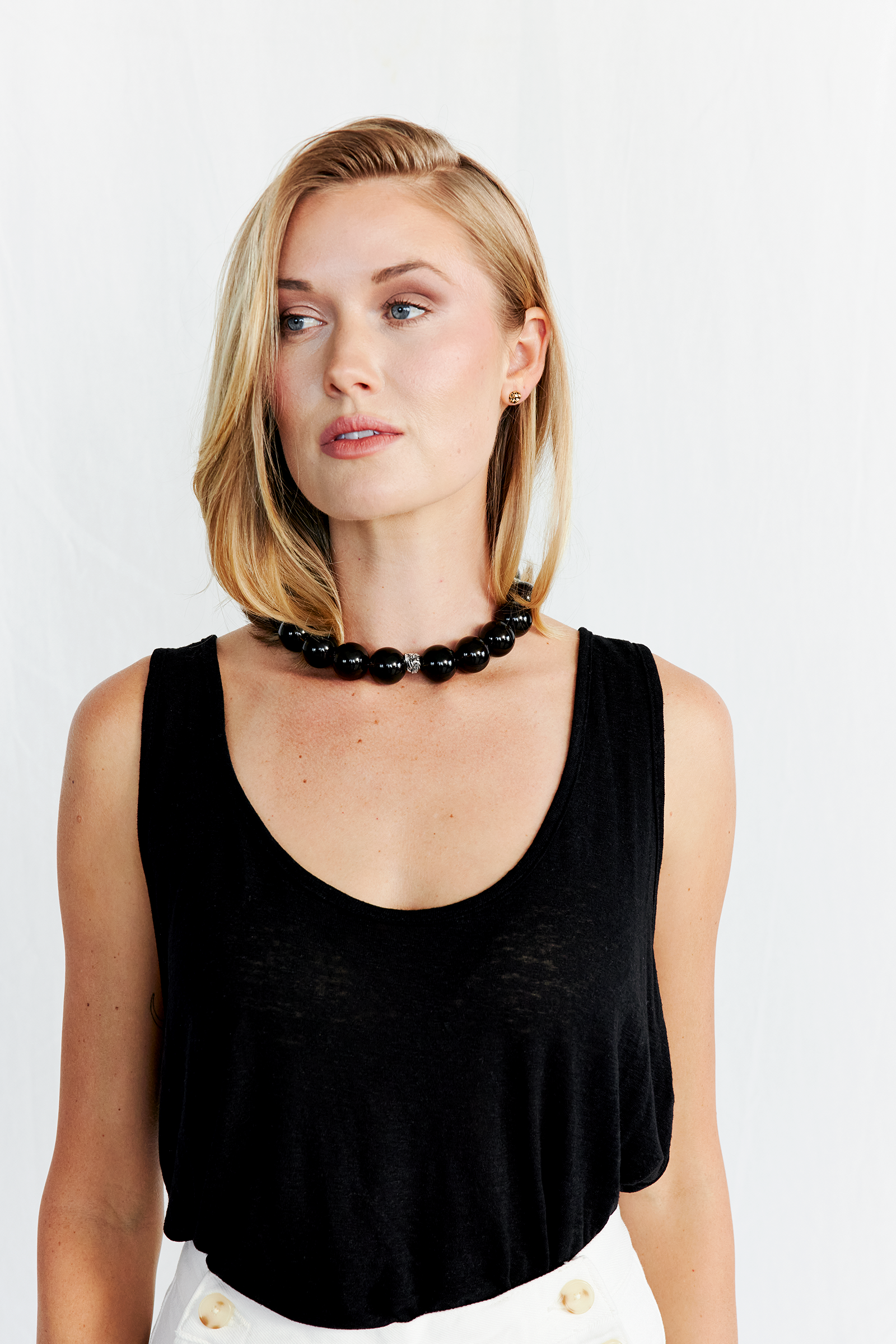 A woman with shoulder-length blonde hair wearing a black sleeveless top, a black beaded necklace, pearl earrings, and white pants.