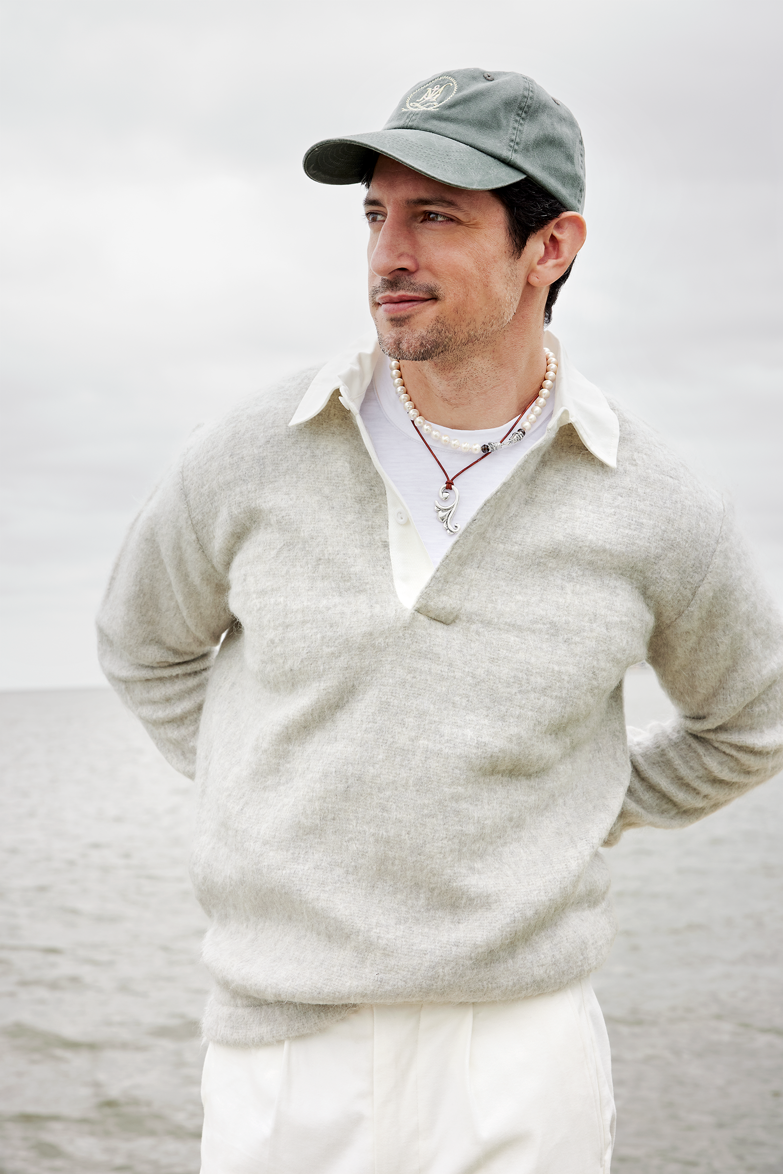 A man wearing a gray cap, white collared shirt, light gray sweater, and white pants stands outdoors near a body of water. He has a pearl necklace and a silver pendant necklace, with hands behind his back, looking to the side with a slight smile.