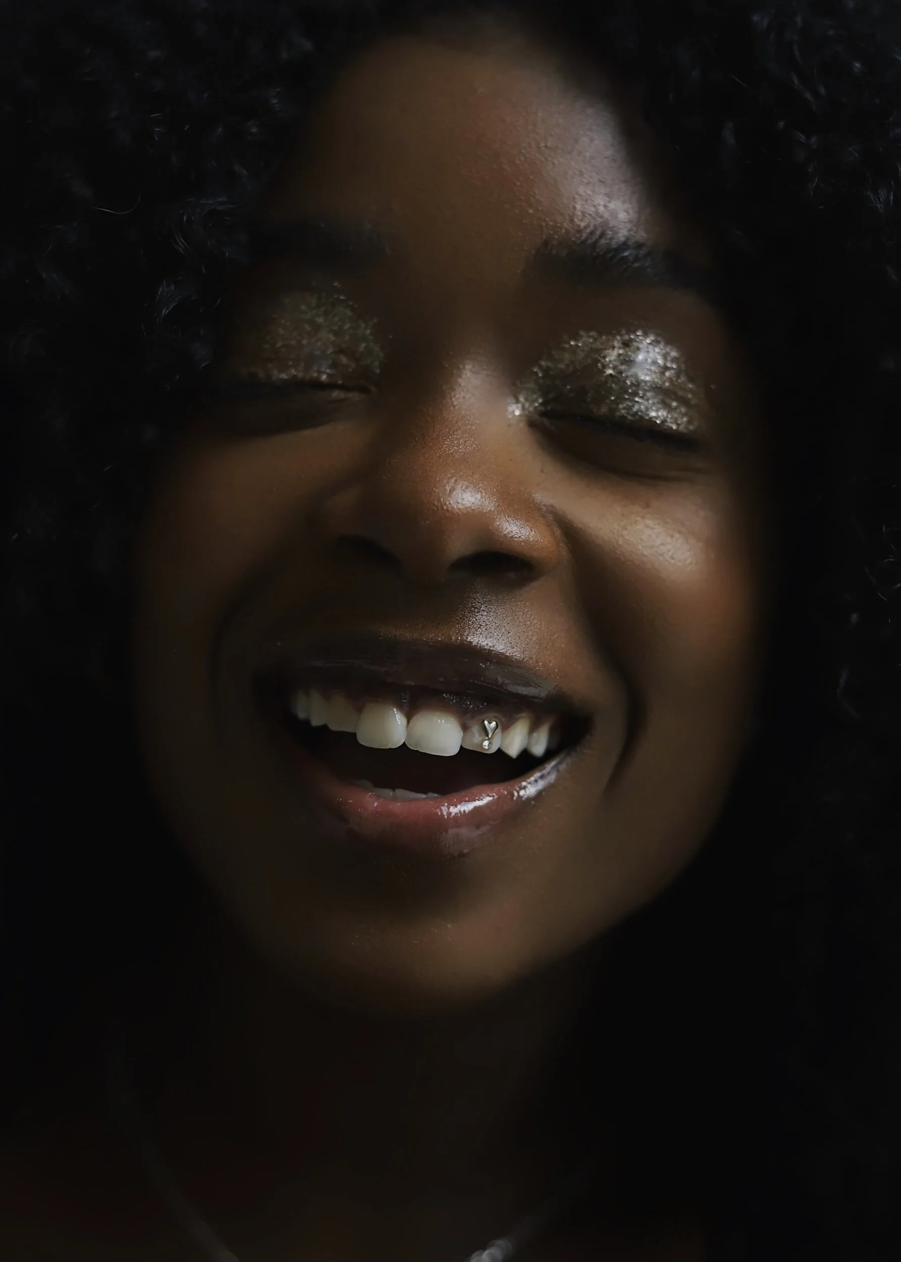 Close-up of a woman with dark curly hair smiling with eyes closed, showing tooth gem , with shiny golden eyeshadow on her eyelids.