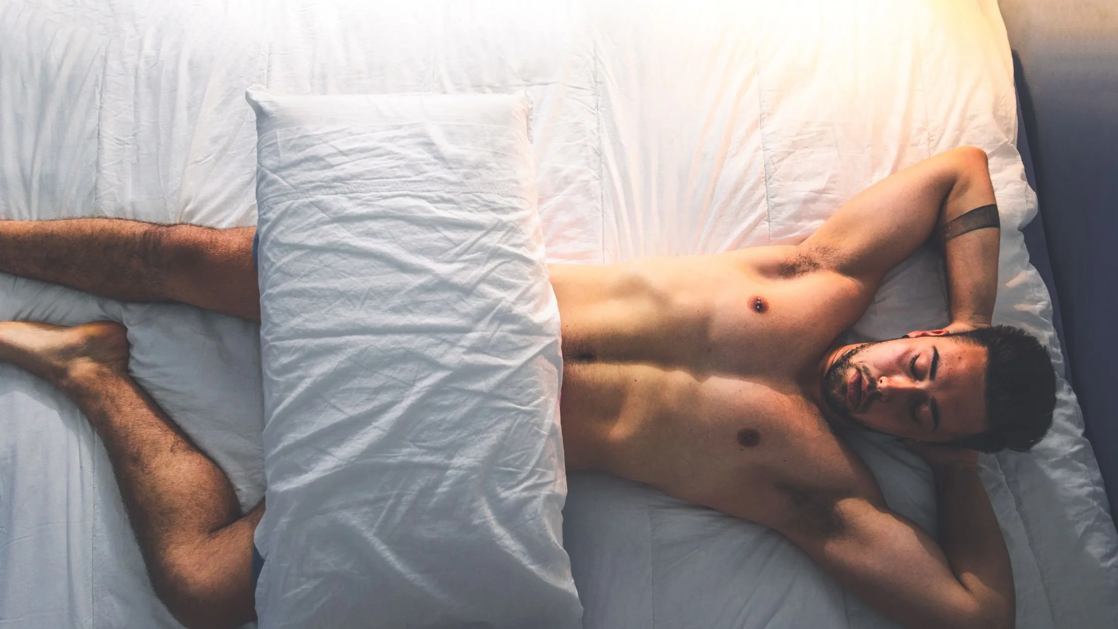 Naked man lying in bed with his eyes closed and a pillow covering his groin