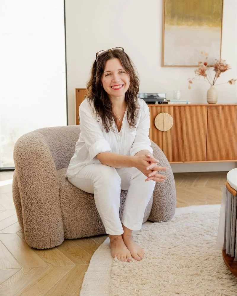 sam sitting on a cream boucle chair, dressed in white , hands clasped, glasses on head and smiling