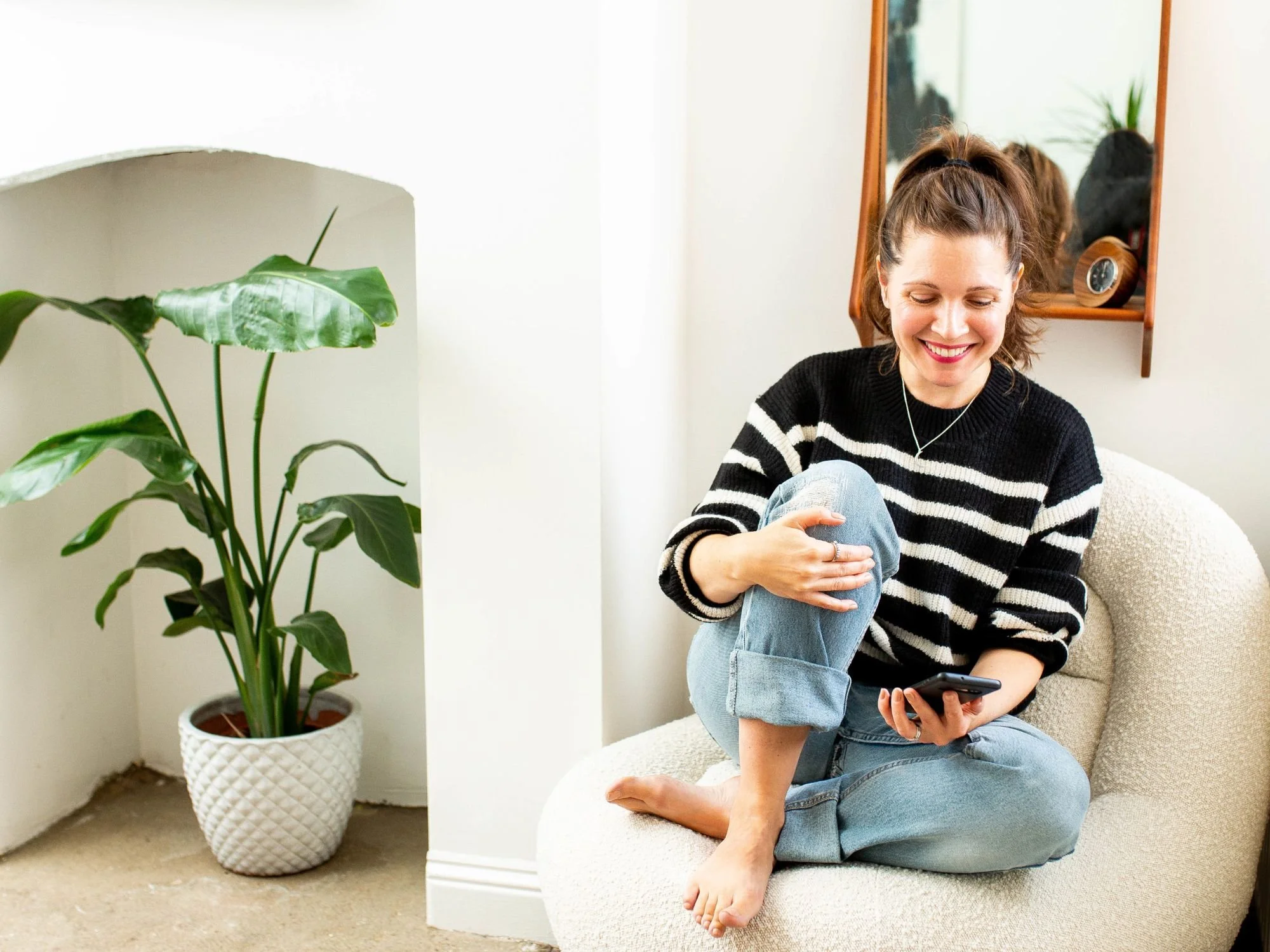 Sam ferguson sitting on a white boucle aormchair and looking at her phone, indoor green plant to her right.