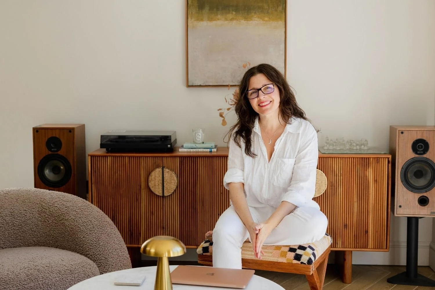sam sitting on a patchwork living room stool, dressed in white , hands clasped, glasses on and smiling