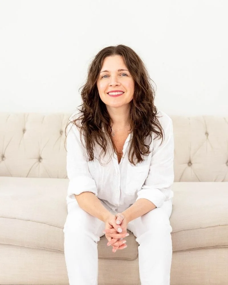 sam sitting on a cream sofa, dressed in white , hands clasped and smiling