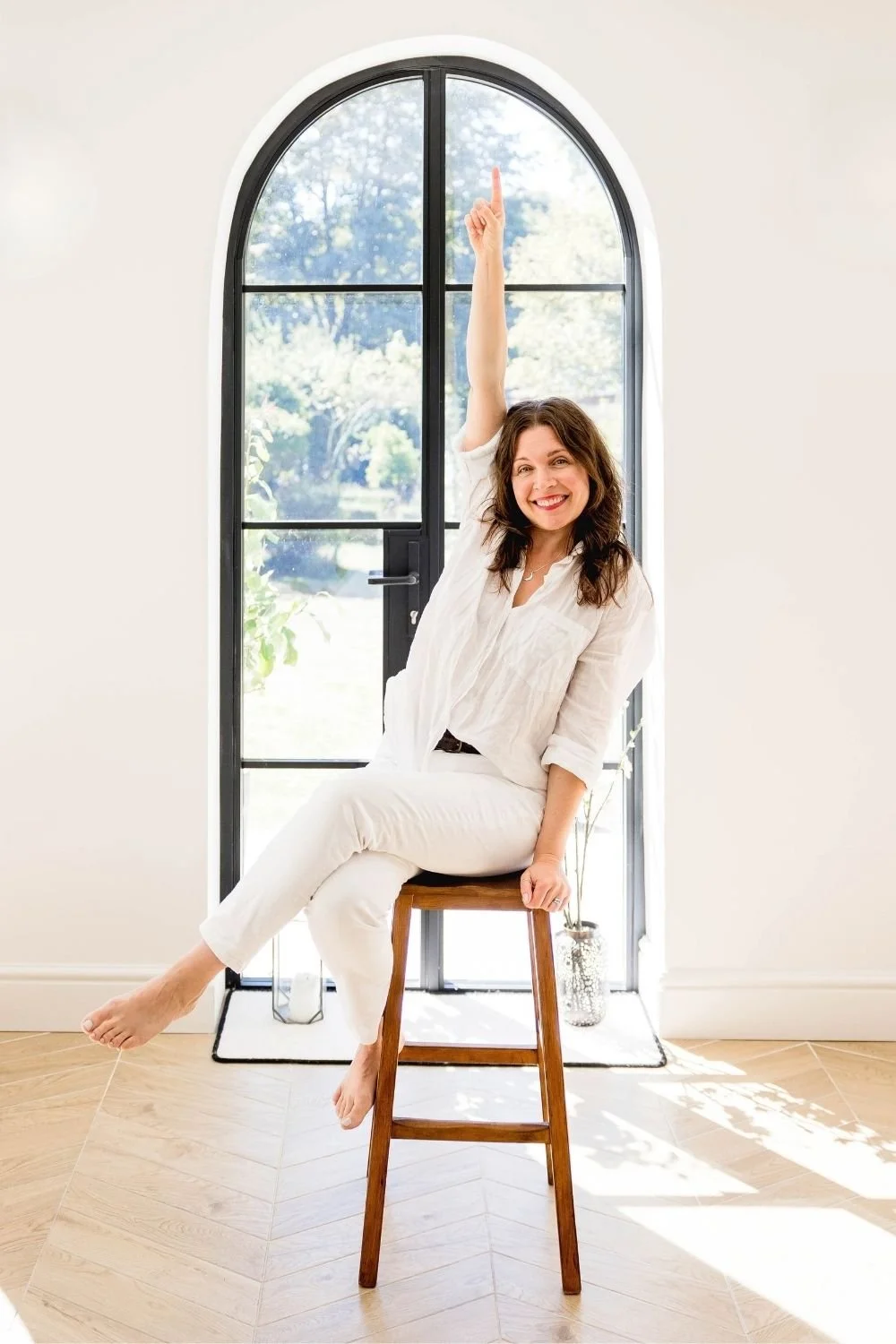 Sam on stool and pointing upwards smiling, dressed in white jeans and white linen shirt