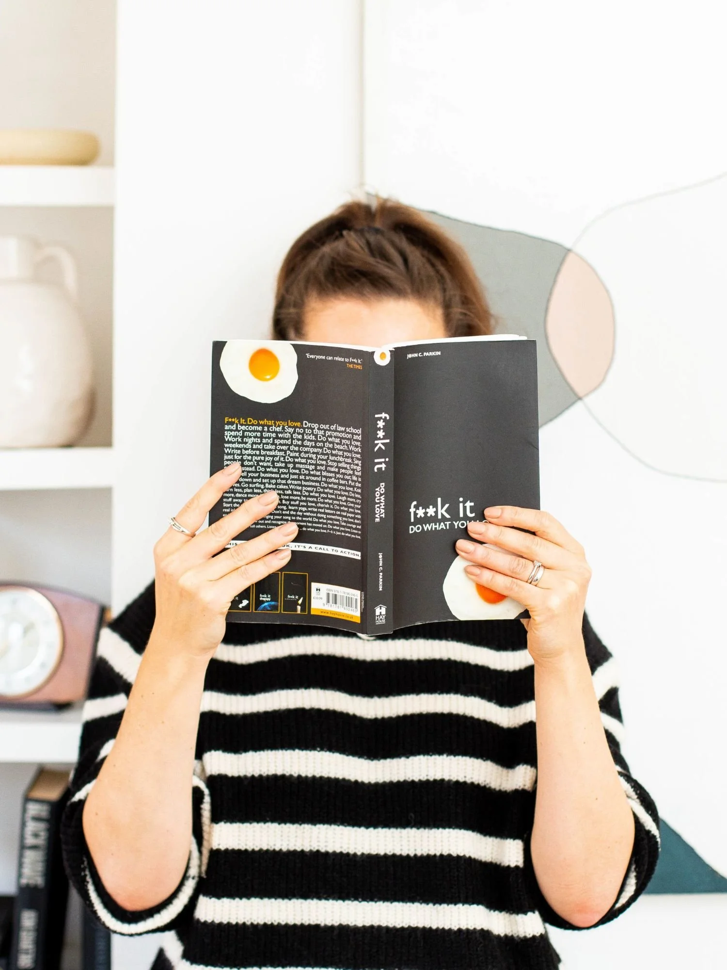 woman holding book covering her face, in stripy jumper