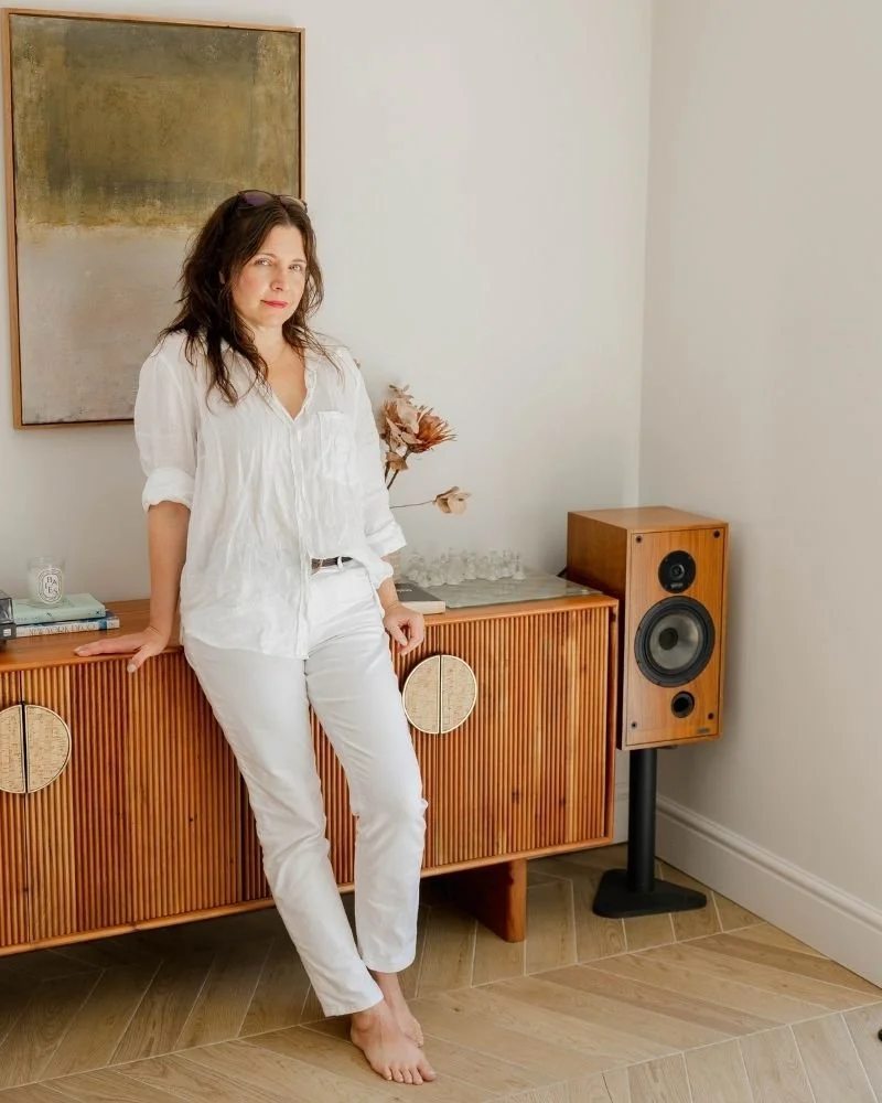 sam leaning against a living room cabinet, dressed in white , glasses on head and smiling