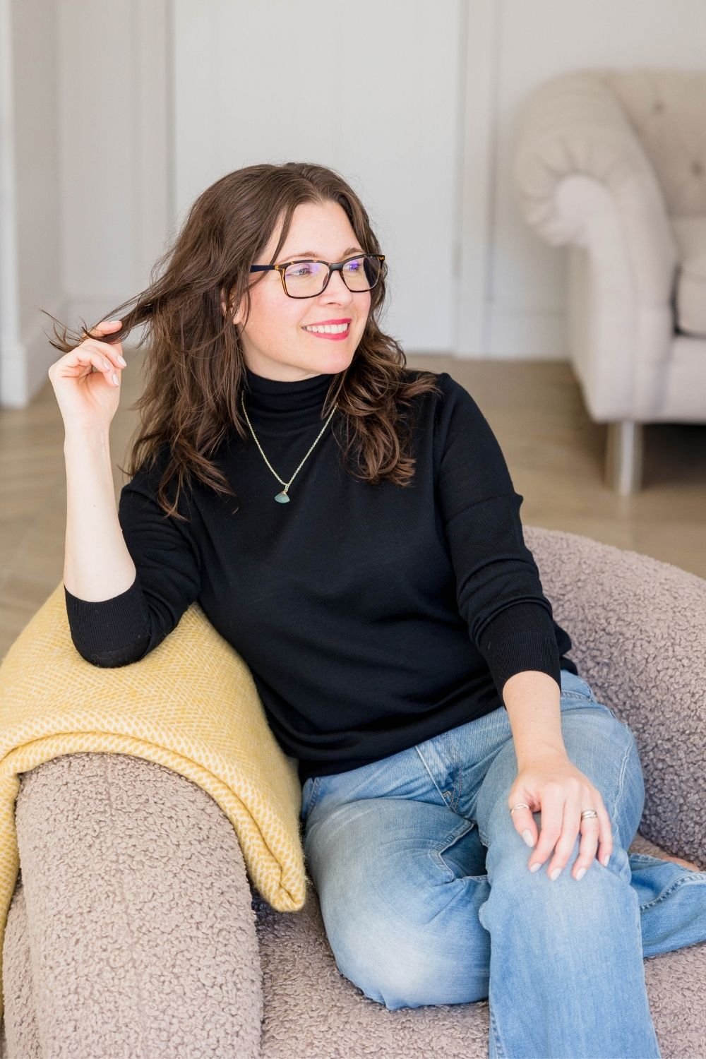 woman wearing black jumper and jeans, sitting on beige boucle chair, holding strand of hair and looking off to the side