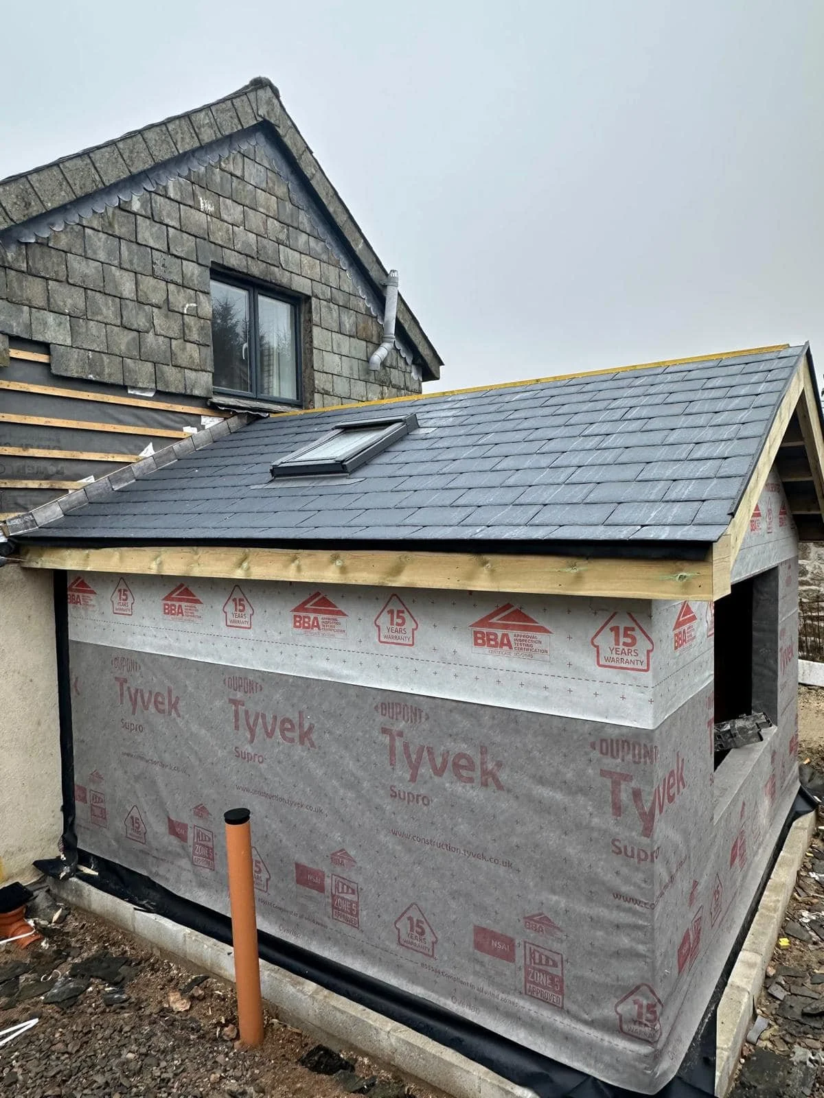 Interior view of a room under renovation with exposed wall studs, debris on the floor, a window letting in sunlight, and construction materials and wires hanging from the ceiling.