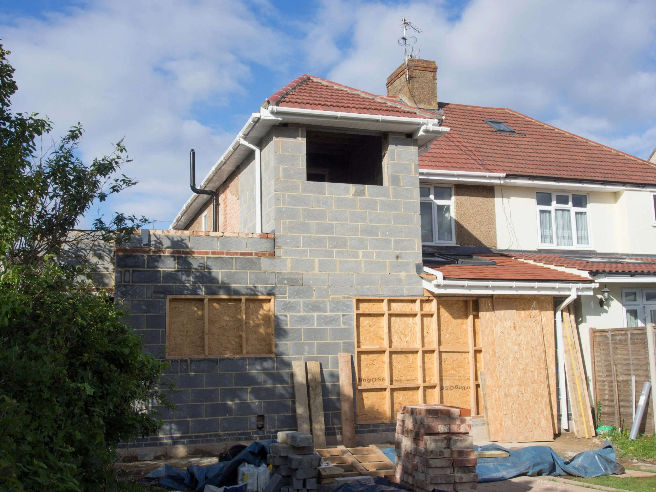 A house undergoing extension or renovation, with new brickwork and wood framing on an addition, construction materials like bricks, wood, and tools in the front yard, and partly cloudy sky above.