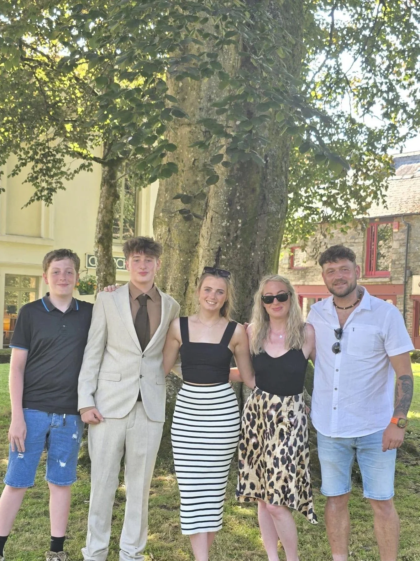 A group of five people standing outdoors in front of a large tree, smiling at the camera. Trees and buildings are visible in the background.