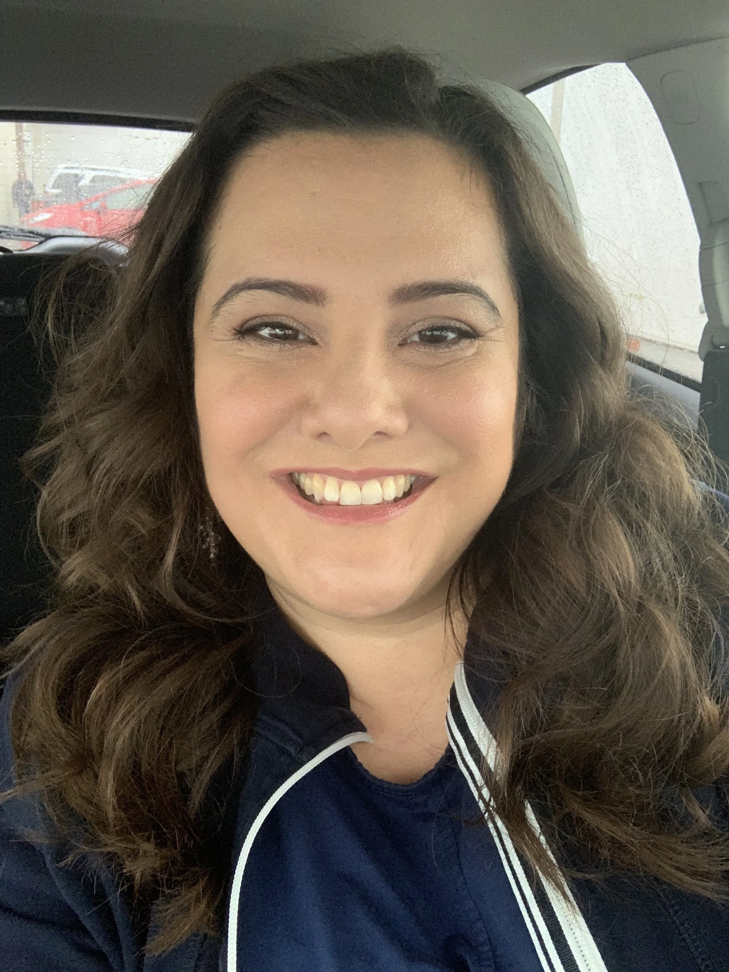 Medium skinned woman with long dark brown wavy hair wearing a navy blue blouse with white trim and smiling.