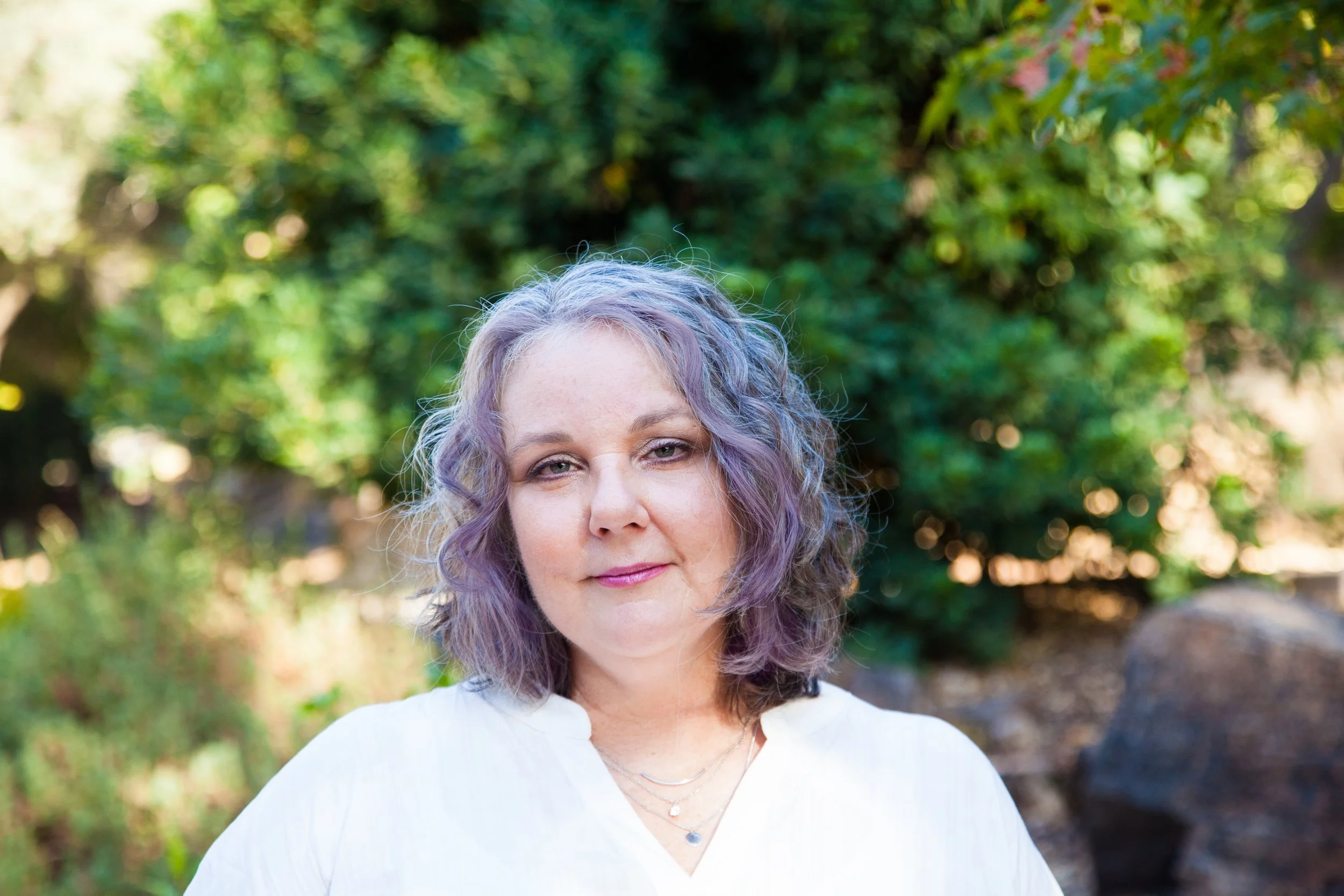 Light skinned femme with purple and blue wavy hair smiling wearing a White blouse and wearing chain necklaces.