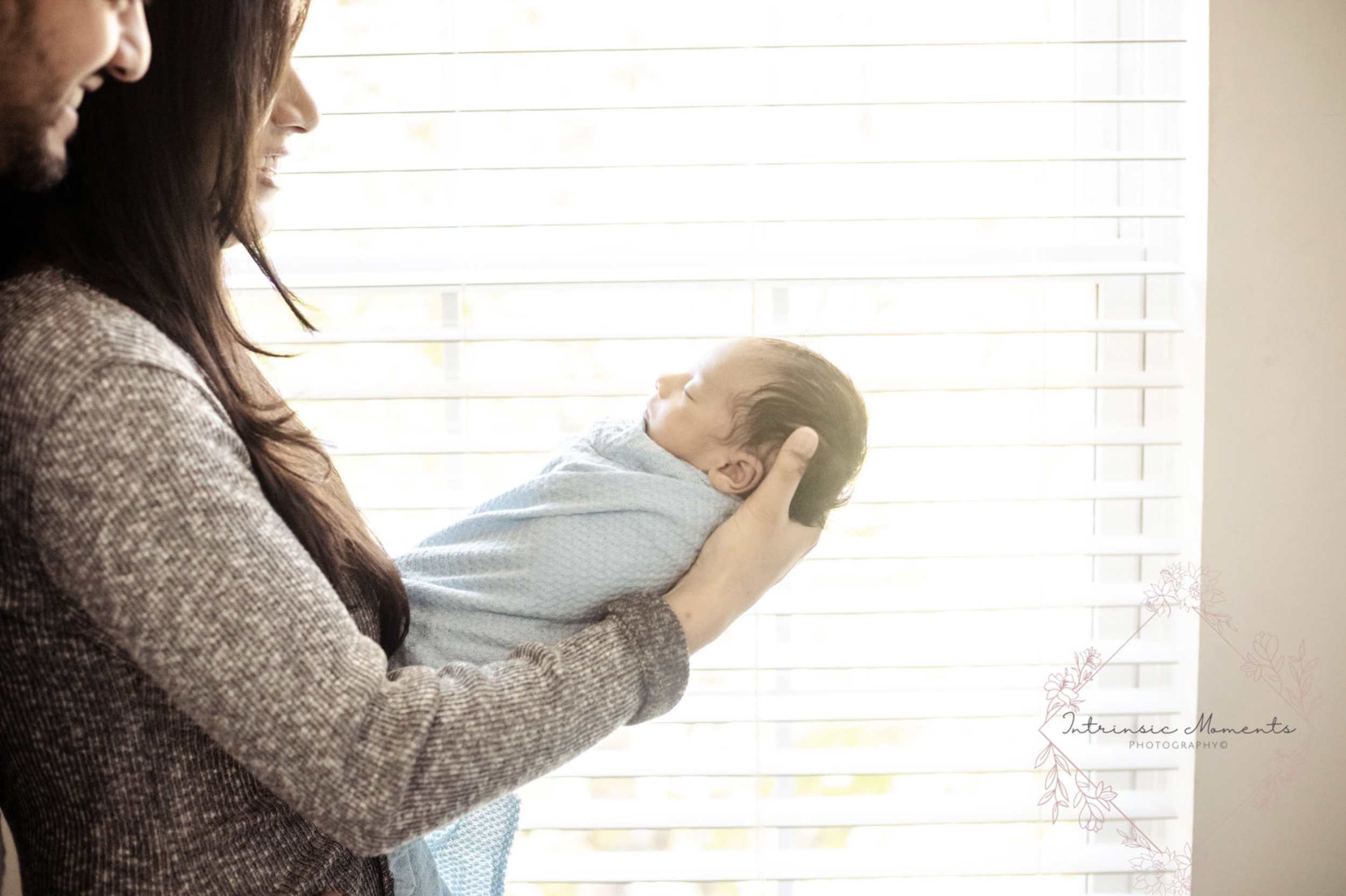 Parents holding newborn baby wrapped in a blue blanket by a window with blinds.