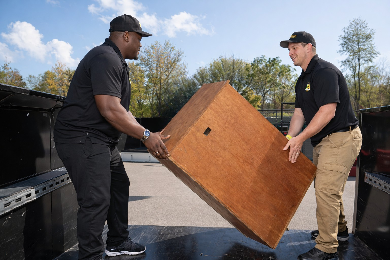 Two men in black shirts and khaki pants lifting a wooden cabinet into a truck on a sunny day with trees in the background.