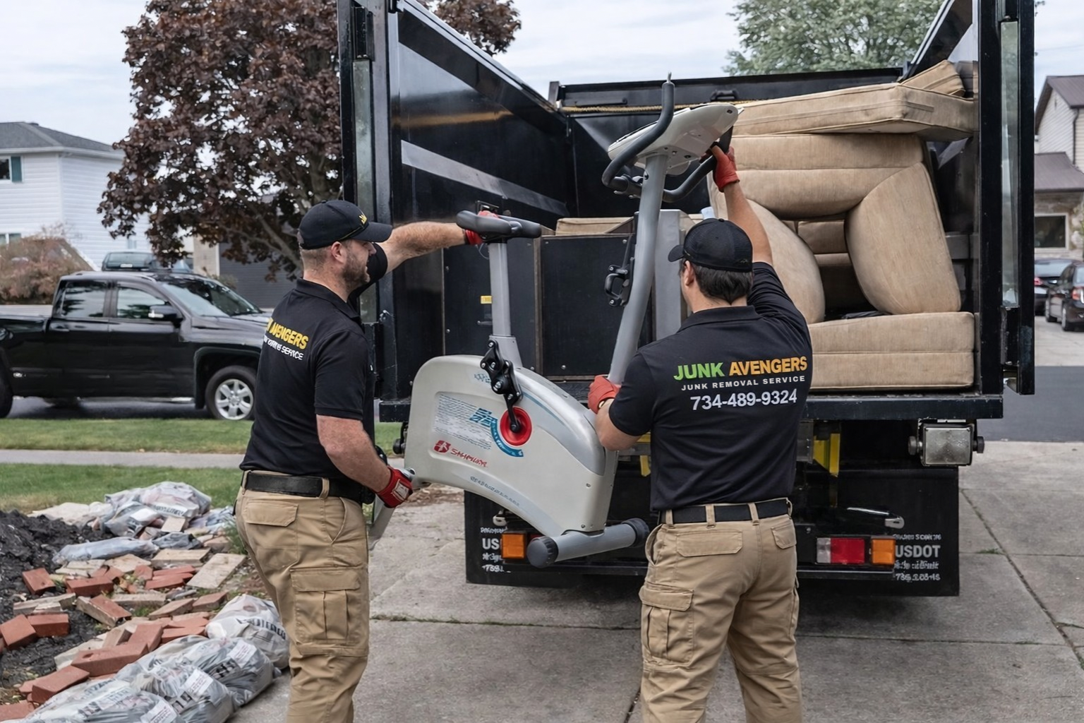 Two workers wearing black shirts with 'Junk Avengers' logo loading furniture into a trash truck. One worker is holding a scooter, and the other is placing a sofa inside the truck. Piles of debris and bricks are on the ground nearby.