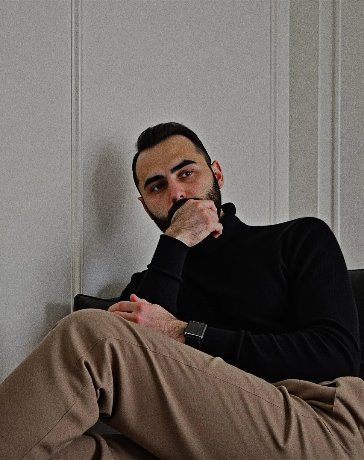 A man with a beard and dark hair, wearing a black turtleneck and tan pants, sitting with his legs crossed against a white paneled wall. He rests his chin on his hand and looks directly at the camera.