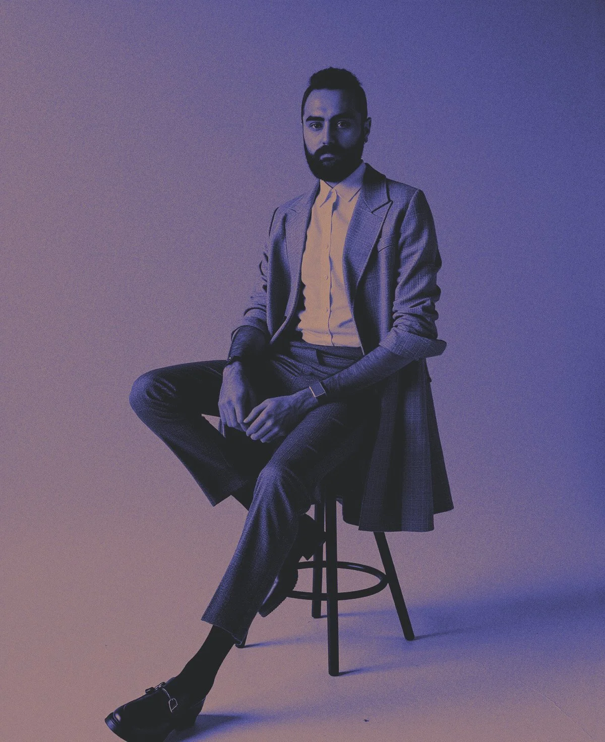 Man sitting on a stool in a gray suit, white shirt, and black shoes against a gradient background.