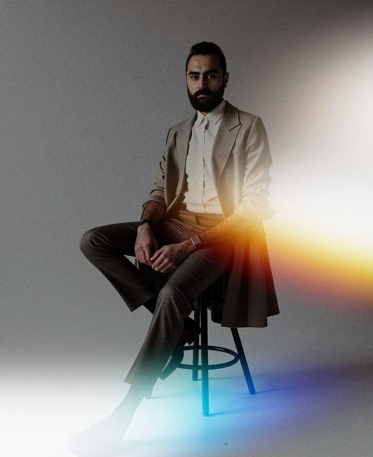 A man with a beard in business attire sitting on a stool with a dark gray background. There is a colorful light flare in the image.