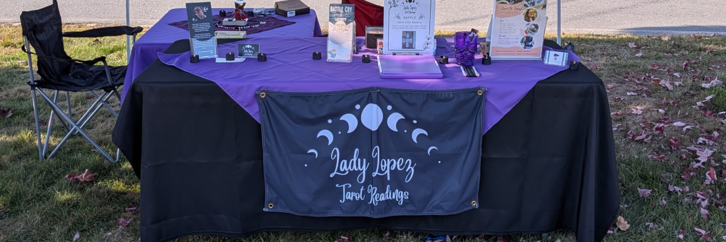 Image of a vendor table with a black and purple table cloth, and a black banner that says Lady Lopez Tarot