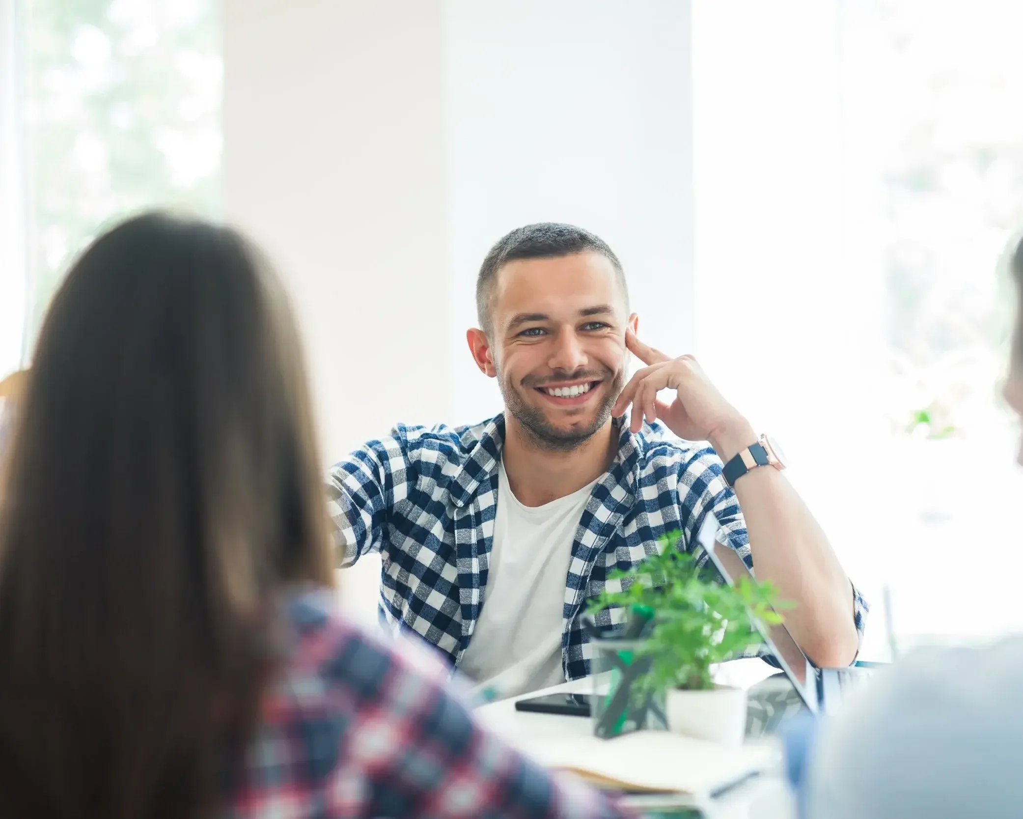 A smiling man in a checkered shirt sitting at a table with two women and a green potted plant in a bright room.