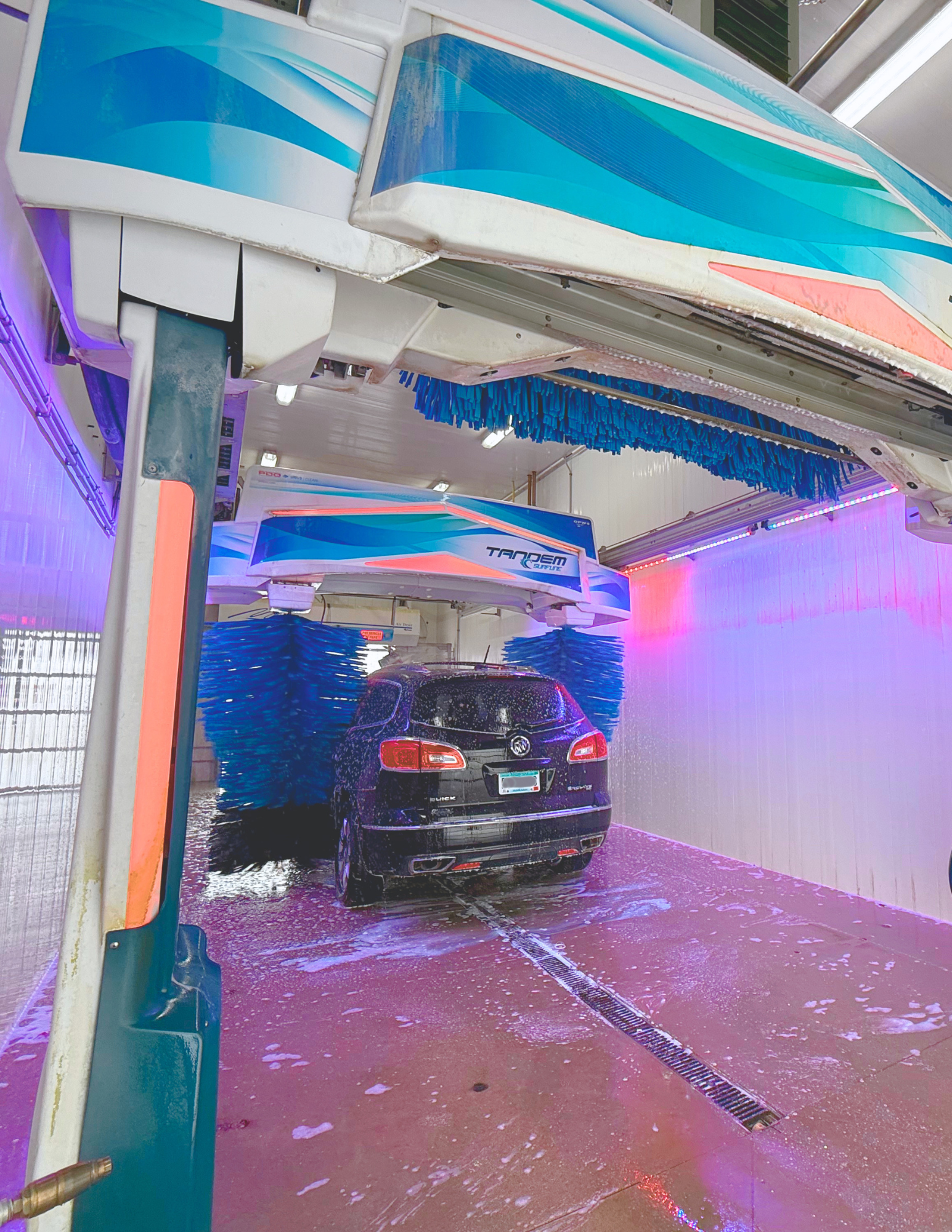 An indoor car wash with a dark SUV being cleaned by rotating brushes and water spray, illuminated by colorful neon lights.