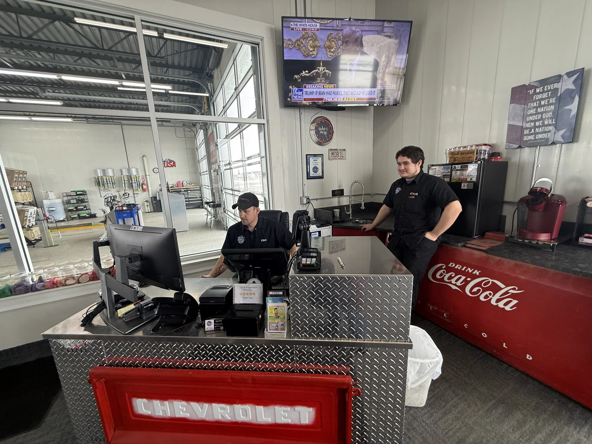 Two employees working at the counter of a car parts store. One is seated and working on a computer, while the other stands nearby. The counter is metallic with a red Chevrolet sign. Behind them is a Coca-Cola cooler, and a television is mounted on the wall. The store has a garage door and various car parts and tools visible in the background.
