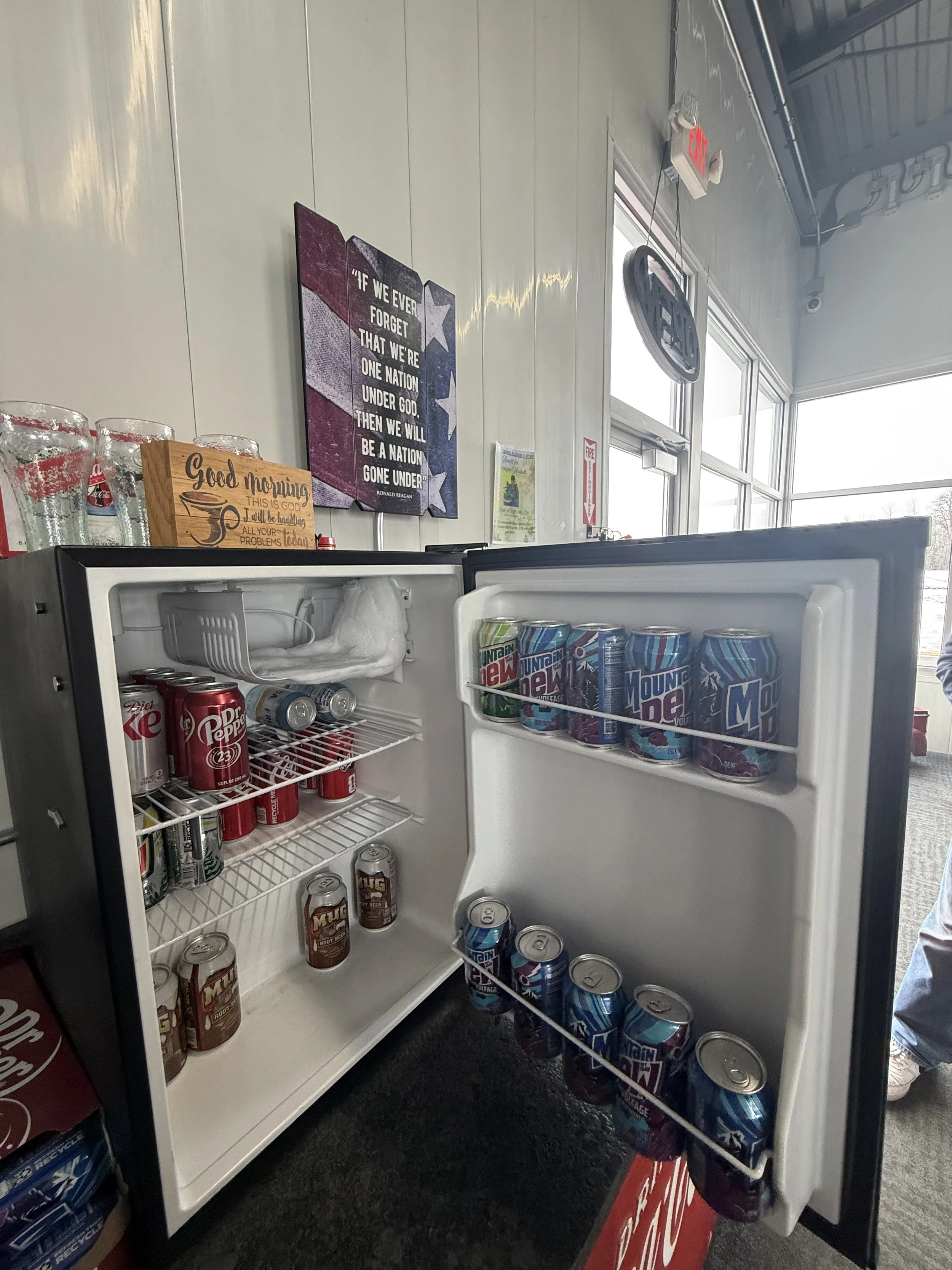 Open mini refrigerator with cans of Mountain Dew, Dr. Pepper, and Mug Root Beer inside, located in a room with white walls, a sign that reads 'Good morning, this is God. All your problems today, I will be handling them for you,' and a wall decoration with an American flag-inspired quote.