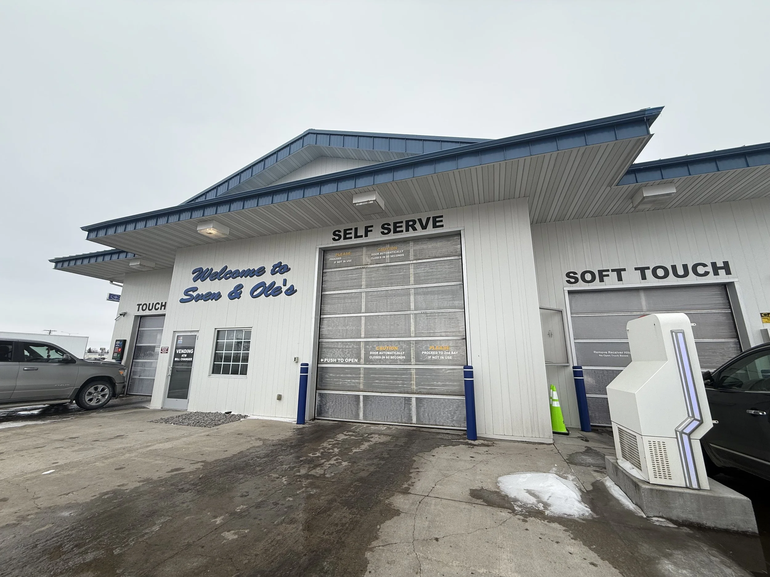 Front view of a self-serve car wash station with two bays, one labeled 'TOUCH' and the other 'SOFT TOUCH', featuring large garage-style doors, a white building exterior with blue accents, a car parked on the left, and a bright, cloudy sky.