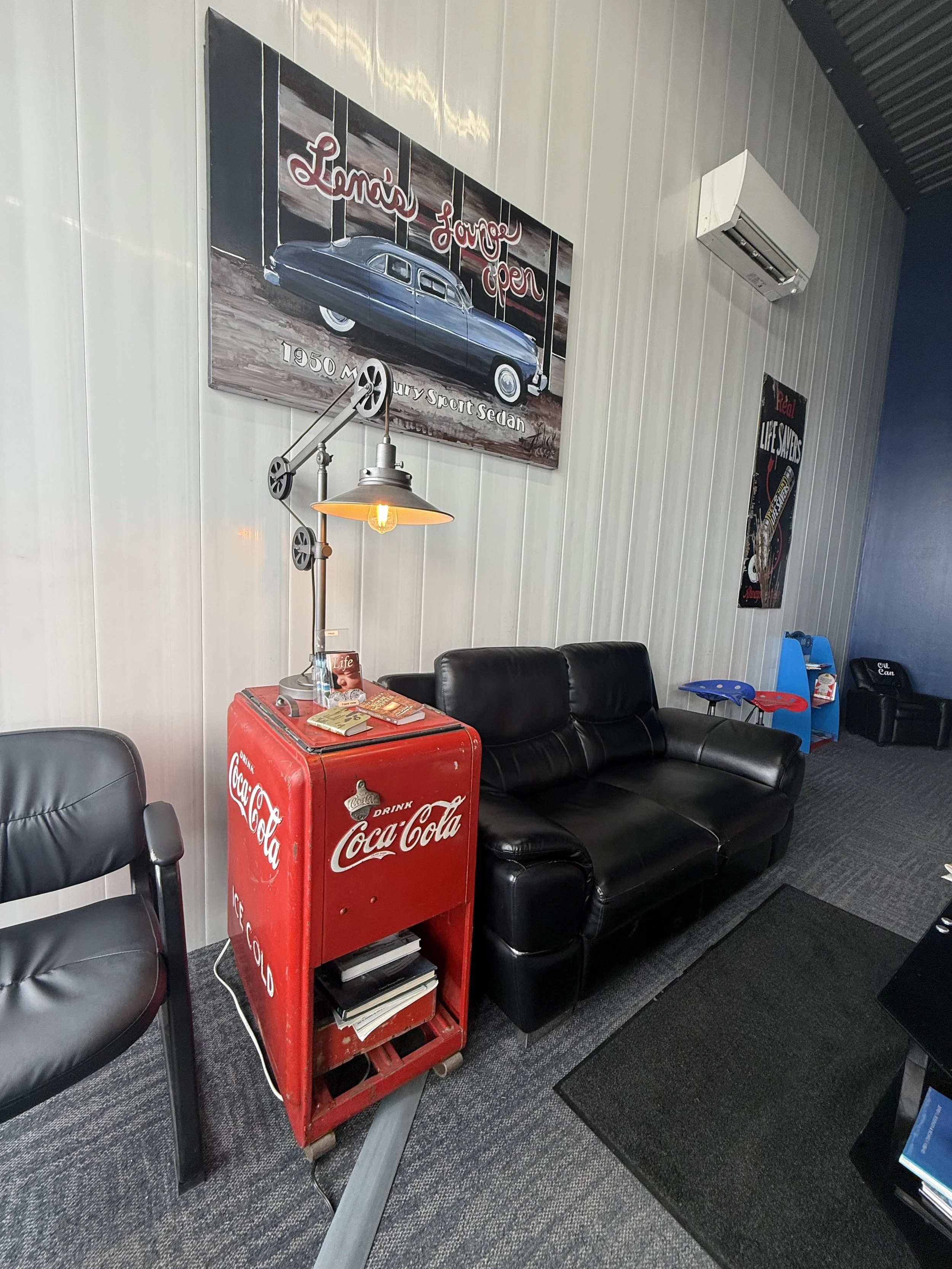 Vintage-themed waiting area with a black leather sofa, a Coca-Cola branded red cooler used as a side table with magazines, a black armchair, and posters on the wall including a car and tattoo themes. There is a wall-mounted air conditioner and a hanging industrial-style lamp.
